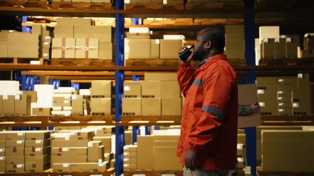 Black warehouse operator using portable walkie-talkie radio in distribution hub. Staff coordinates shipment details, parcel tracking and cargo handling with two-way transmitter in depot.