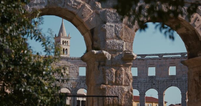 View of the bell tower of the Church of Saint Anthony framed by the ancient stone arches of the Pula Arena Roman amphitheater in Pula Croatia.