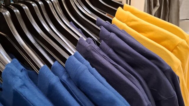 Close-up high angle view of colorful cotton t-shirts hanging on black plastic hangers in a retail clothing store. The neat rows of blue, navy, and yellow garments create an organized commercial displa