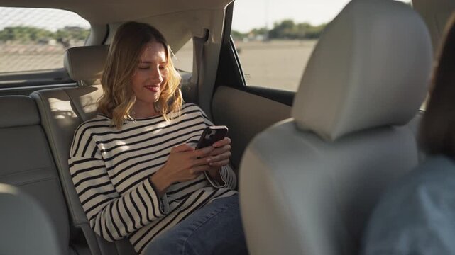 Smiling woman in striped sweater sitting in back seat of car, holding smartphone, tapping with both hands and looking at screen. Passenger texting on mobile phone and looking through window.