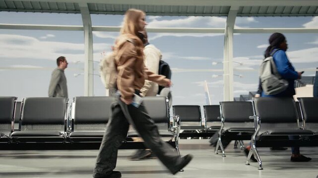 Multiethnic men and women commuting in airport transport hub overlooked by runway control tower. Multiracial travelers walking through metal chairs inside flight departure terminal, camera A