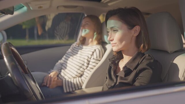 Two beautiful women sitting in car front seats, driver in black shirt looking ahead with focused expression, passenger in striped sweater leaning back and smiling. Sunlight patterns through windshield