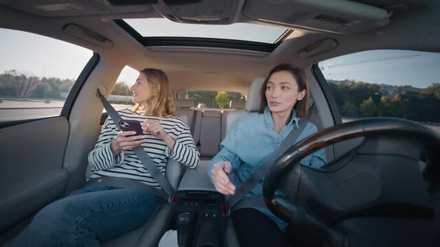 Two Caucasian women sitting in car front seats talking and gesturing while driving in daylight. Female driver explaining direction to passenger using smartphone for navigation during trip.