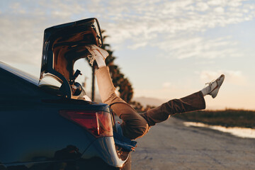 Naklejka premium Woman lifestyle, car, road, beach, sunset, sunglasses, travel, casual, outdoors, she leans on open trunk, warm golden sunlight, relaxed stance, seaside breeze, cinematic sky
