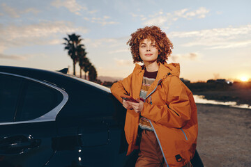 Naklejka premium Woman lifestyle outdoors, sunset light, car exterior, orange coat, curly hair, portrait pose, street evening, calm mood, hands adjusting jacket, natural colors