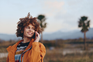 Obraz premium Outdoor woman lifestyle portrait, smiling young woman with curly hair in an orange jacket, gently touching hair outdoors in a rural field under warm evening light, softly blurred trees