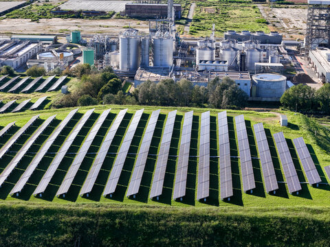Aerial view of solar panels on a green hillside with an industrial plant and storage tanks in the background Crescentino, Piedmont, Italy.