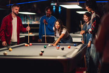 Fototapeta premium Young diverse friends enjoying a night out, playing pool together at a bar, laughing and having fun