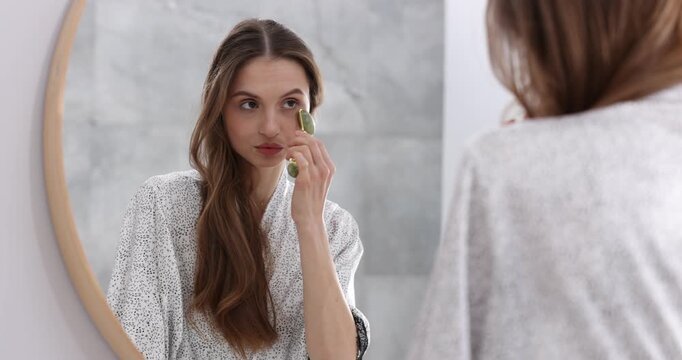 Beautiful woman doing facial self massage with roller near mirror indoors, selective focus