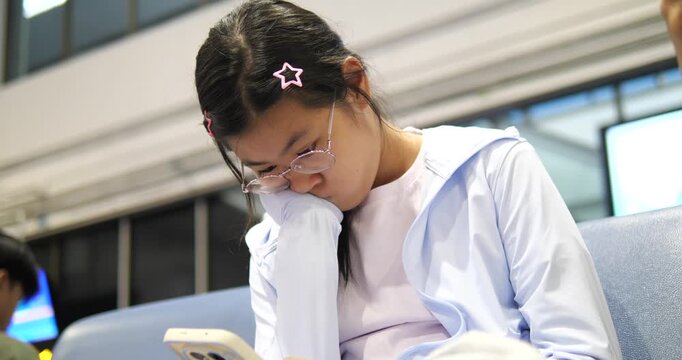 Cinematic close-up of a young Asian girl with glasses using a smartphone while waiting at an airport terminal, travel lifestyle and digital technology concept