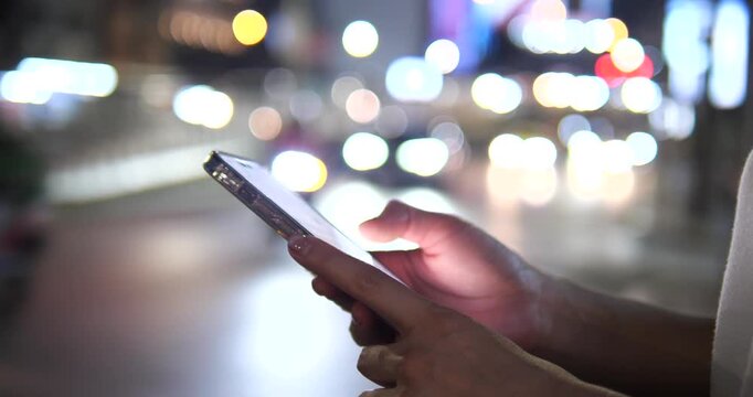 Cinematic close-up of hands using a smartphone on a city street at night, beautiful colorful bokeh background and urban lifestyle concept