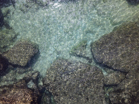 Aerial view of clear turquoise water over submerged dark rocks with sunlight reflections Cascais, Lisbon, Portugal.