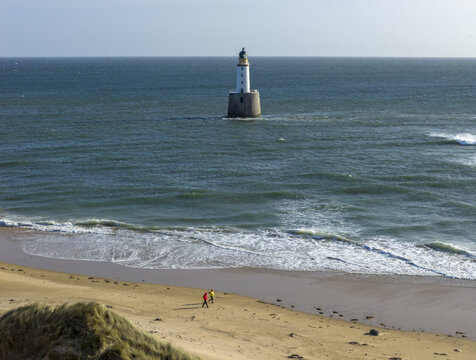 Aerial view of Rattray Head Lighthouse in the North Sea with waves crashing and people walking on the sandy beach in Peterhead, Scotland, United Kingdom.