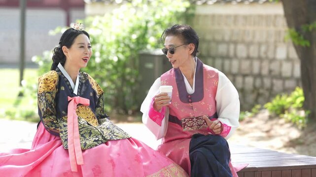 Two women in traditional Korean attire sit on a bench. One smiles warmly, the other laughs gently with a cup. They converse, dressed in bright, patterned hanboks. Background features stone walls