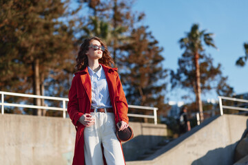 Obraz premium Candid woman outdoors wearing a red coat, sunny day, relaxed posture, soft breeze, natural colors, railing backdrop, fashion forward mood, confident expression