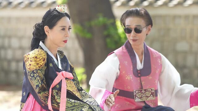 Two women in traditional Korean attire sit near a stone wall. One wears a patterned dark robe with pink accents. The other dons a vibrant pink vest over white. They engage in conversation