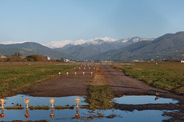 Naklejka premium A dirt road cuts through a vast field toward distant snowcapped mountains beneath a clear blue sky, creating a tranquil, expansive rural landscape with peaceful solitude