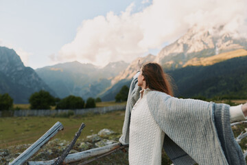 Obraz premium Woman in a chunky knit shawl stands with outstretched arms in a mountain meadow, embracing outdoor lifestyle and serene natural beauty.
