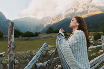 Obraz premium Woman wrapped in a gray knit blanket stands outdoors, gazing upward with mountains in the background, evoking a cozy, serene lifestyle moment in nature.