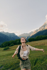Obraz premium Joyful woman hiking in a green meadow with mountains in the background, wearing a light jacket and backpack. Outdoor adventure and wellness. Embracing nature Outdoors.