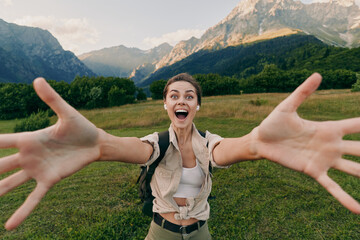 Obraz premium Cheerful woman outdoors, reaching toward the camera with arms wide, sunlit field and mountains behind, casual outfit, joyful lifestyle moment, natural colors