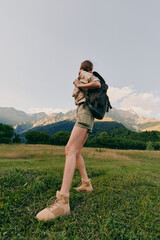 Obraz premium Woman hiking outdoors with a child on her back, backpack and boots in a meadow beneath mountains, nature, adventure, and family lifestyle.