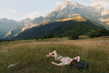 Obraz premium A young woman lies on a sunlit meadow with rugged mountains in the background, resting beside a backpack, conveying relaxation and a sense of outdoor freedom.