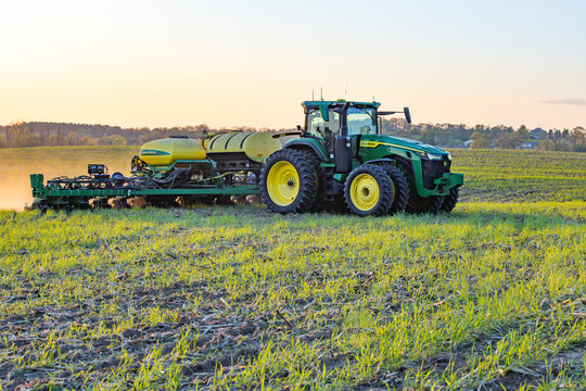 A John Deere 310 R8 tractor pulling a John Deere ExactEmerge corn planter with liquid fertilizer tanks through a field with cover crops at sunset.