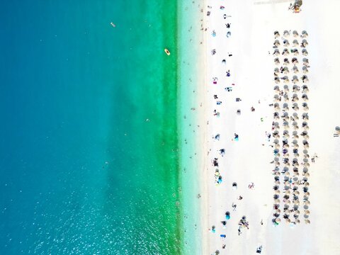 Aerial view of a sandy beach with turquoise water, white sand, and rows of sun umbrellas with people swimming and sunbathing.