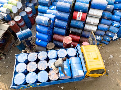 Aerial view of workers loading blue metal barrels onto a yellow truck next to large stacks of industrial drums in Dhaka, Dhaka Division, Bangladesh.