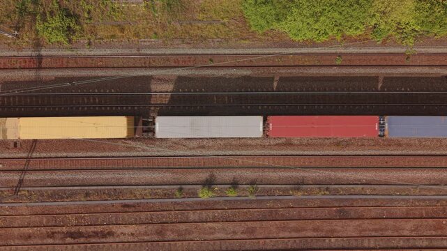 Vertical aerial drone footage looking straight down at cargo freight train transporting colorful shipping containers along railway tracks in the United Kingdom.