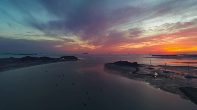 Drone pullback parallax shot from Jebudo tidal flats gradually reveals Jeongok Marina with yachts and anchored trawlers against vivid sunset sky