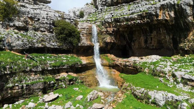 Scenic drone footage of a waterfall dropping beside cave-like rock formations and green terrain in Qattin Azar, Baskinta, Lebanon.
