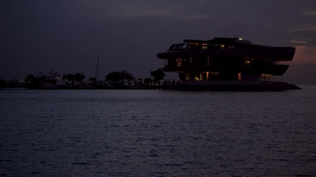 Calm nighttime waterscape featuring a striking modern seaside pavilion. The building is silhouetted against a dark sky with glowing windows.