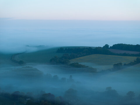 Aerial view of rolling green hills and lush fields shrouded in thick morning mist under a soft dawn sky in Nuffield, Oxfordshire, United Kingdom.