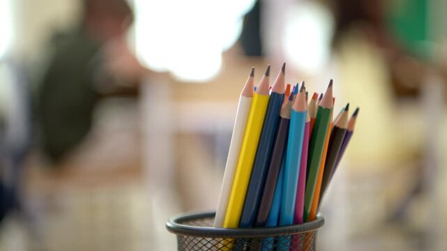 Close up of colorful pencils in a container inside a classroom with blurred students in the background