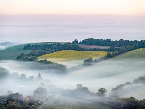 Aerial view of rolling green hills and lush fields partially covered by thick morning mist under a soft dawn sky Nuffield, Oxfordshire, United Kingdom.