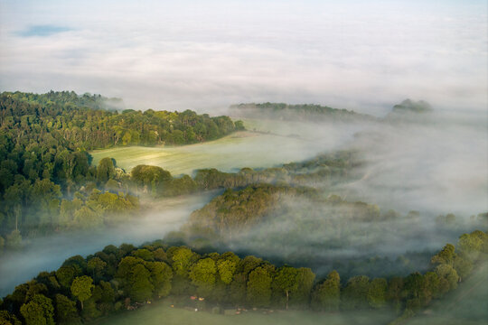 Aerial view of rolling green hills and dense woodlands partially covered by thick morning mist under a soft light Nuffield, Oxfordshire, United Kingdom.