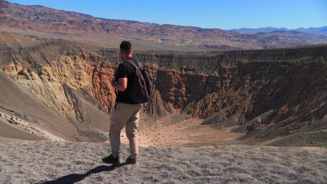 Male hiker with backpack walks along edge of massive Ubehebe volcanic crater in Death Valley National Park, taking in stunning panoramic views of colorful layered rock walls in orange.