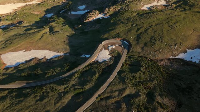 Aerial footage of Beartooth Highway in Montana showcasing a serpentine mountain road curving through alpine terrain with melting snow patches and green tundra at golden hour.