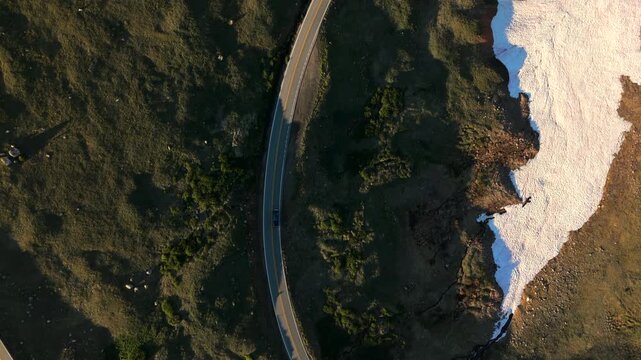 Aerial drone footage follows lone vehicle driving along winding Beartooth Highway in Montana as golden sunset light illuminates rugged alpine landscape with melting snow patches contrasting against.
