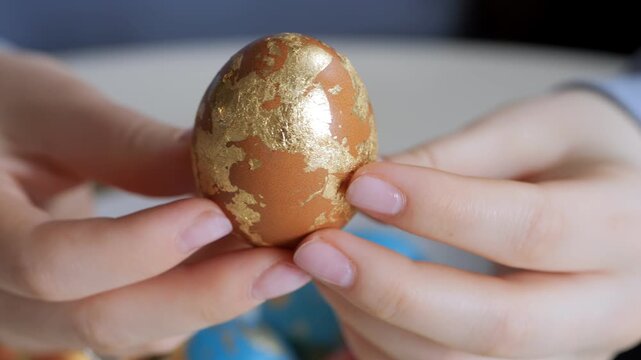 Hands hold decorated egg with golden leaf pattern. Egg has brown base color. Background shows colorful eggs. Close-up shot of intricate design. Focus on craftsmanship and detail