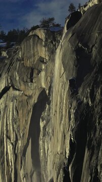 Vertical Screen: Yosemite National Park Firefall phenomenon with water streaming down massive granite rock face, sunlight illuminating the cliff with pine trees silhouetted against blue sky