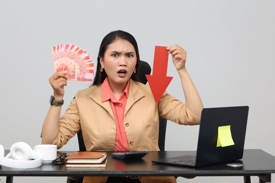 A frustrated young female employee holds a paper arrow pointing down indicating a decrease in income while holding rupiah cash banknotes, working at an office desk with a laptop PC.