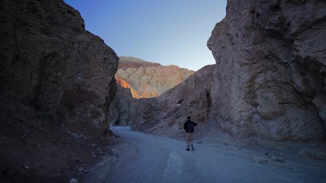A lone hiker with backpack walks along a gravel path through a dramatic narrow canyon. Towering rock walls frame the scene as golden sunlit mountains glow in the distance. Golden canyon, Death Valley.