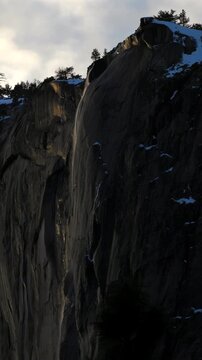 Vertical Screen: Yosemite National Park Firefall phenomenon with water streaming down dramatic granite cliff face covered in snow patches and silhouetted pine trees against cloudy winter sky