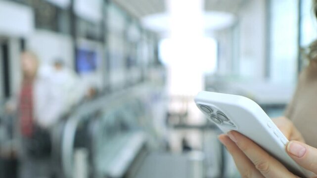 Hand holds smartphone in public transit area. Escalator and people blurred in background. Modern indoor setting with soft lighting. Focus on phone use during commute