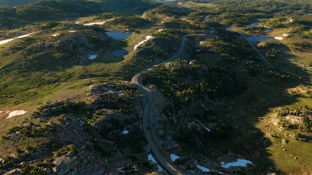 Beartooth Highway in Montana curves through stunning alpine terrain with scattered lakes and snow patches. Aerial view reveals winding road crossing rocky highlands dotted with evergreen trees.