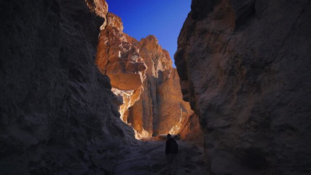Lone hiker walks through narrow desert slot Golden canyon, Death Valley, with towering rock walls. Golden sunlight illuminates orange sandstone cliffs against deep blue sky creating dramatic contrast.