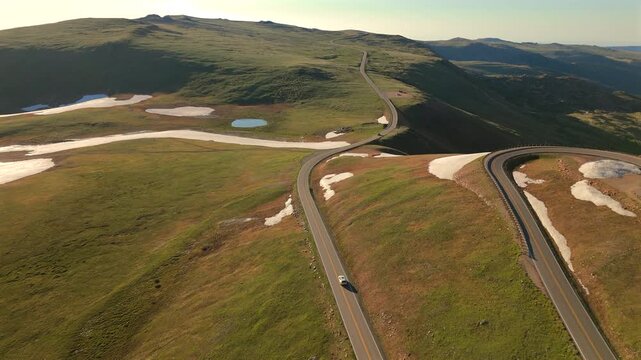 Drone footage captures Beartooth Highway in Montana winding through lush green alpine meadows with melting snow patches and pristine mountain lakes during golden hour light.